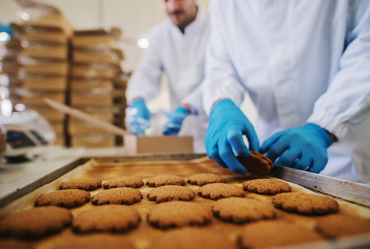 close-up-tray-full-fresh-baked-cookies-food-factory-blurred-picture-two-male-employees-sterile-clothes-packing-cookies-background