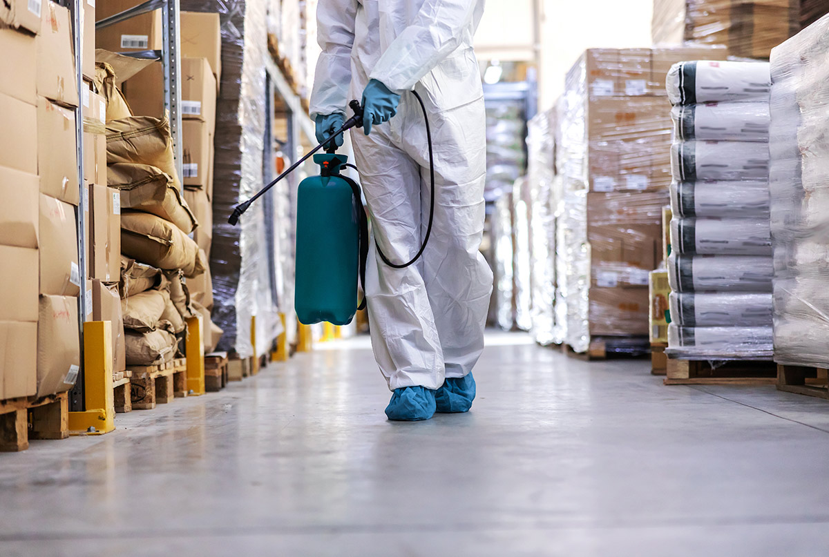 worker-sterile-uniform-with-rubber-gloves-holding-sprayer-with-disinfectant-spraying-around-warehouse-corona-outbreak-concept